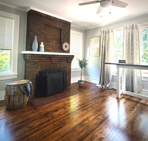 A room with a brick fireplace, wooden flooring, a ceiling fan, and a window with curtains. - 10 Gorgeous, contemporary Apt on Historic Earle St (Greenville)