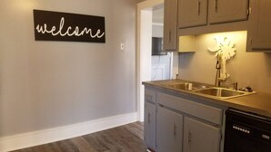 A kitchen with grey cabinets, a stainless steel sink, and a dishwasher. A 'welcome' sign is on the wall. - 10 Historic Earle with Rustic Charm (Greenville)