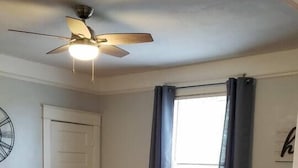 Una sala de estar con un sofá gris oscuro, un reposapiés a juego, un ventilador de techo, un reloj en la pared y una ventana con cortinas.