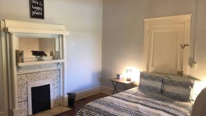 Un dormitorio con chimenea, una cama con ropa de cama estampada, una mesita de noche con lámpara y una estantería de pared.