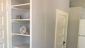 Una habitación con una estantería blanca empotrada, un cubo de basura negro, una puerta blanca y un refrigerador blanco.
