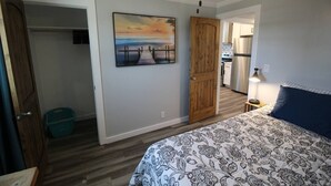 Ein Schlafzimmer mit Bett, Deckenventilator, Holztür, einer Küchenzeile und einem Gemälde an der Wand.
