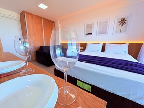 A hotel room with a bed, a bedside table with a wine glass and a plate, a wooden nightstand, and a view of the outside. - Studio com vista Mar em Maceió – Max Rev (Maceió)