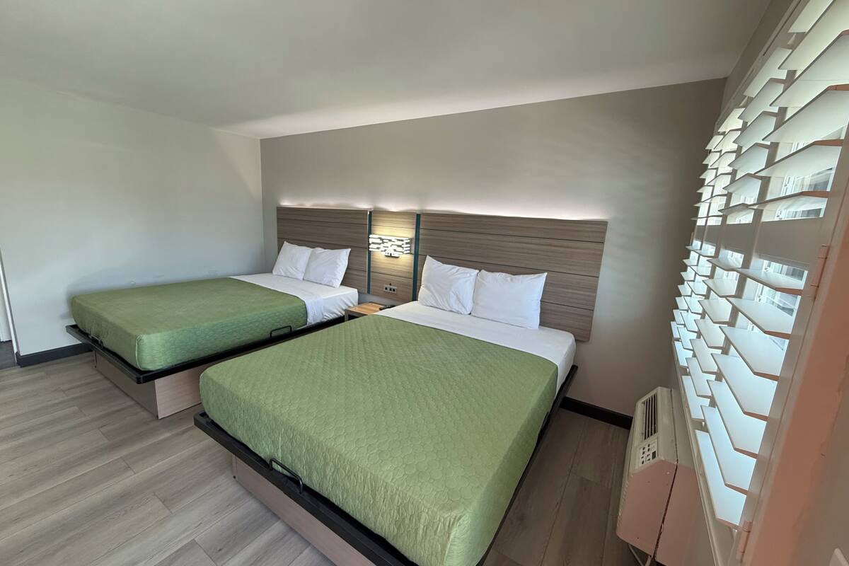A hotel room with two beds, a wooden headboard, a wall-mounted light, and a built-in shelving unit.