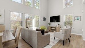 Un salon spacieux avec de hauts plafonds, de grandes fenêtres, un téléviseur à écran plat, un fauteuil vert et une table à manger en bois avec des chaises.