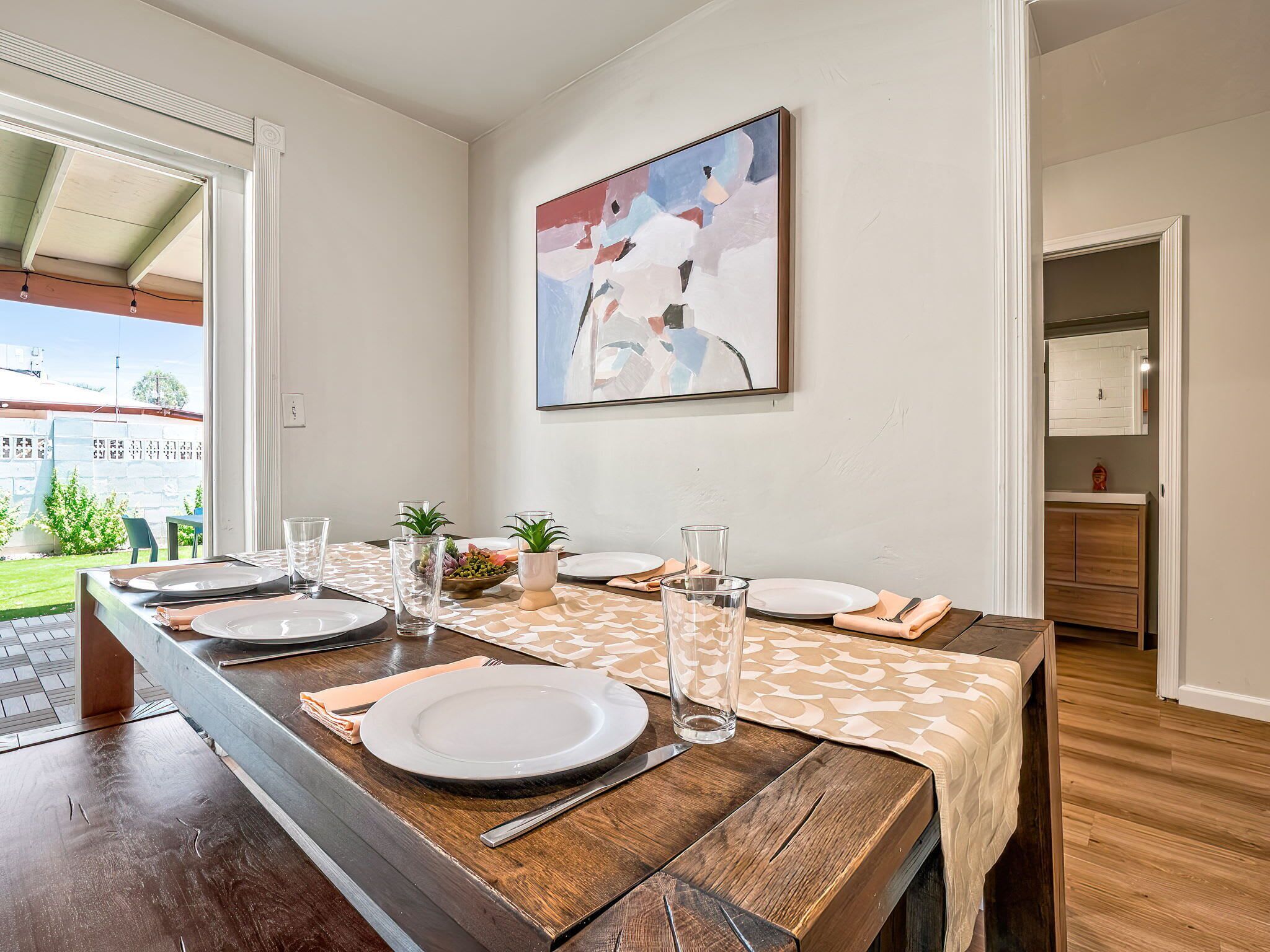 Een eetruimte met een lange houten tafel, gedekt voor vier personen, een schilderij aan de muur en uitzicht op het buitenleven.