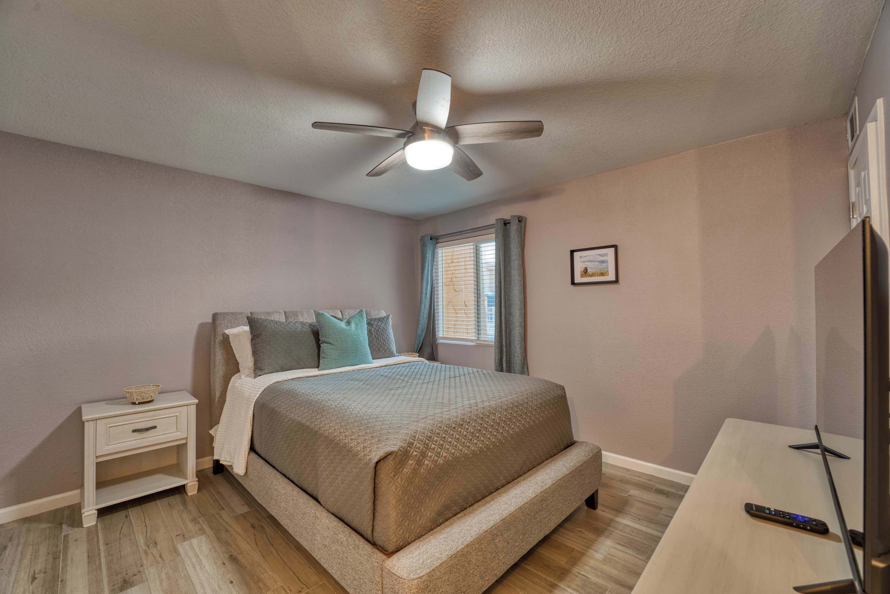 A bedroom with a bed, nightstand, ceiling fan, window with curtains, and a TV stand.