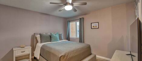 A bedroom with a bed, nightstand, ceiling fan, window with curtains, and a TV stand.