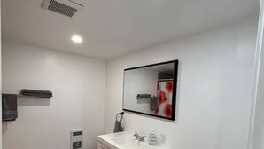 Une salle de bain avec un lavabo blanc, des toilettes, un miroir et une grille d’aération au plafond.