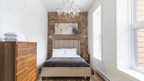 A bedroom with a brick wall, a bed, a wooden dresser, a chandelier, and a large window.