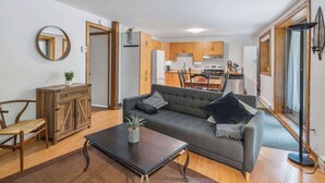 Una sala de estar con sofá, mesa de centro y aparador de madera. Se observa una cocina al fondo con muebles de madera y un comedor.