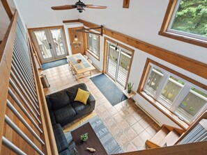 A spacious loft-style room with a high ceiling, a dining area, a sofa with a cushion, and large windows offering a view of the outdoors.