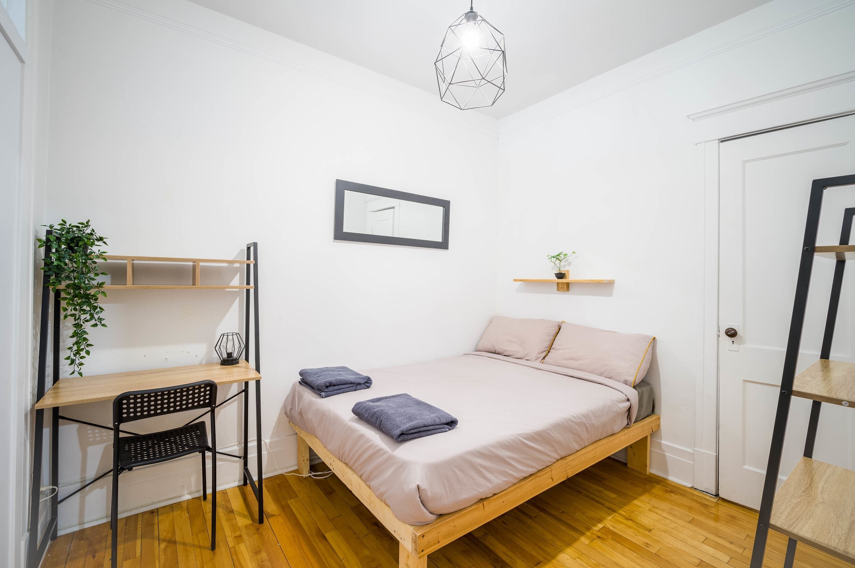 Een kleine, schone kamer met een bed, bureau, stoel, hanglamp en een deur.
