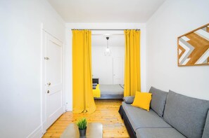 Um quarto com sofá cinzento, cortinas amarelas, chão de madeira, porta branca e cama com cabeceira cinzenta.