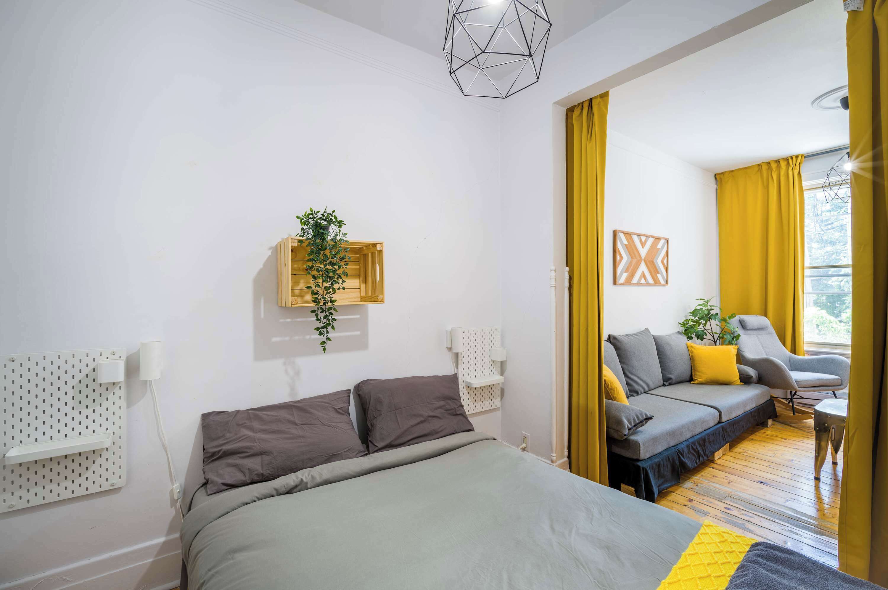A modern hotel room with a bed, a sofa, a small table, a wall-mounted plant, and a geometric ceiling light.