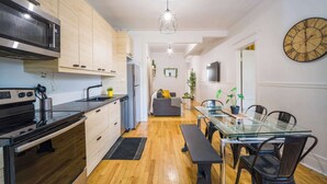 A modern kitchen with a microwave, stove, and wooden cabinets. There is a dining area with a glass table and chairs, and a living room with a sofa and a TV. - Trendy 4BR Montreal Home Perfect for Groups (Montréal)