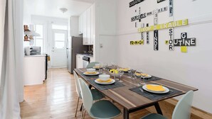 A dining area with a wooden table set for four, white walls, and a kitchen in the background. - Walk to Olympic Stadium |Modern Montreal Stay - apartment (Montréal)