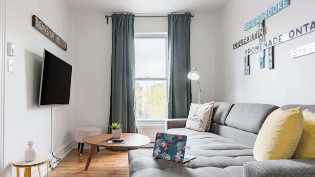 A modern living room with a grey sofa, a flat-screen TV, a round wooden coffee table, and a small potted plant. - Walk to Olympic Stadium |Modern Montreal Stay - apartment (Montréal)
