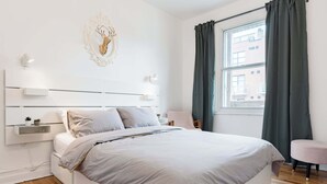 A modern bedroom with a bed, bedside tables, a wall-mounted light, a window with curtains, and a decorative headpiece on the wall. - Walk to Olympic Stadium |Modern Montreal Stay - apartment (Montréal)