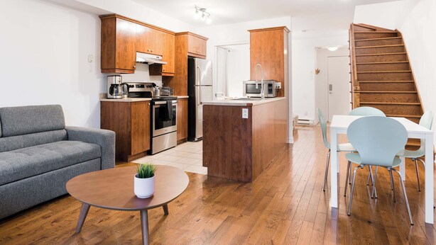 Eine kompakte Küche mit Holzausstattung, einem Kühlschrank, einer Mikrowelle und einer Kaffeemaschine. Es gibt einen Essbereich mit Tisch und Stühlen, und eine Treppe, die zu einer anderen Ebene führt.