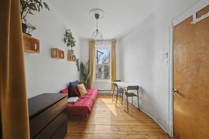 A room with a wooden floor, a red sofa, a small table with chairs, and a window with yellow curtains. - Large 5BR Home  Ideal for Student Groups (Montréal)