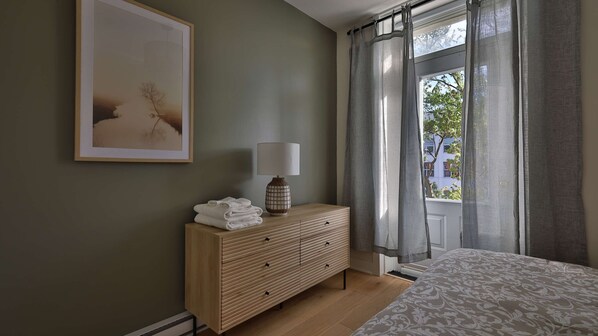 Un dormitorio con una cama, una mesita de noche con una lámpara, un cuadro en la pared y una ventana con cortinas.
