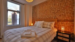 Ein Schlafzimmer mit Ziegelwand, großem Fenster, Bett mit kariertem Überwurf und Nachttisch mit Lampe.