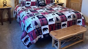 Quarto com uma cama grande, duas luminárias de cabeceira, um painel de madeira na cabeceira, um banquinho de madeira e um ventilador de teto.