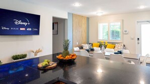 A modern living room with a flat-screen TV displaying Disney+ logos, a dining area with a black countertop, and a seating area with a sofa and yellow cushions.