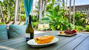 Tavolo apparecchiato all'aperto con una bottiglia di vino, due bicchieri di vino bianco, un piatto con una fetta di papaia, un piatto con uva e una pesca, e una tenda blu sullo sfondo.