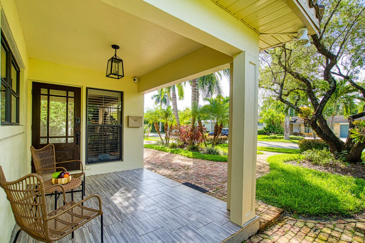 A covered porch with wicker chairs and a table, a brick pathway leading to a garden with palm trees.