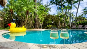 Una piscina con un'anatra gonfiabile gialla, due bicchieri di liquido trasparente e una rigogliosa vegetazione tropicale.