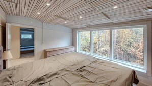 A bedroom with a large bed, a bench, a window with a view of trees, and a wooden ceiling.