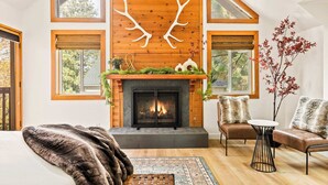Una sala de estar acogedora con chimenea, dos sofás, una mesa de centro y un gran ventanal con vistas a árboles.