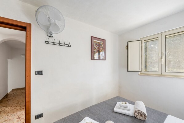 Une pièce comprenant un lit, un ventilateur, un porte-manteau, un tableau encadré et une fenêtre avec des stores.