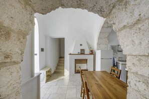 Una habitación con paredes de piedra, una mesa y sillas de madera, una escalera que conduce a otra estancia, y una zona de cocina con nevera y hornilla.