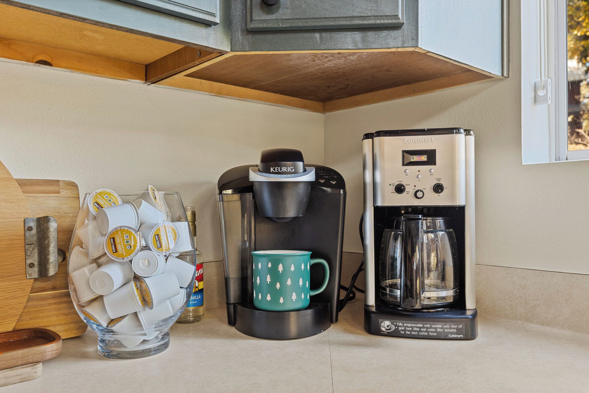 Una cocina con cafetera, una taza y un recipiente con cápsulas de café.