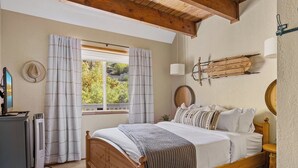 Un dormitorio con cama de madera, una ventana con cortinas, una lámpara de pared y una mesita redonda.