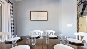 A dining area with a wooden table set for six, white chairs, and a framed quote on the wall.