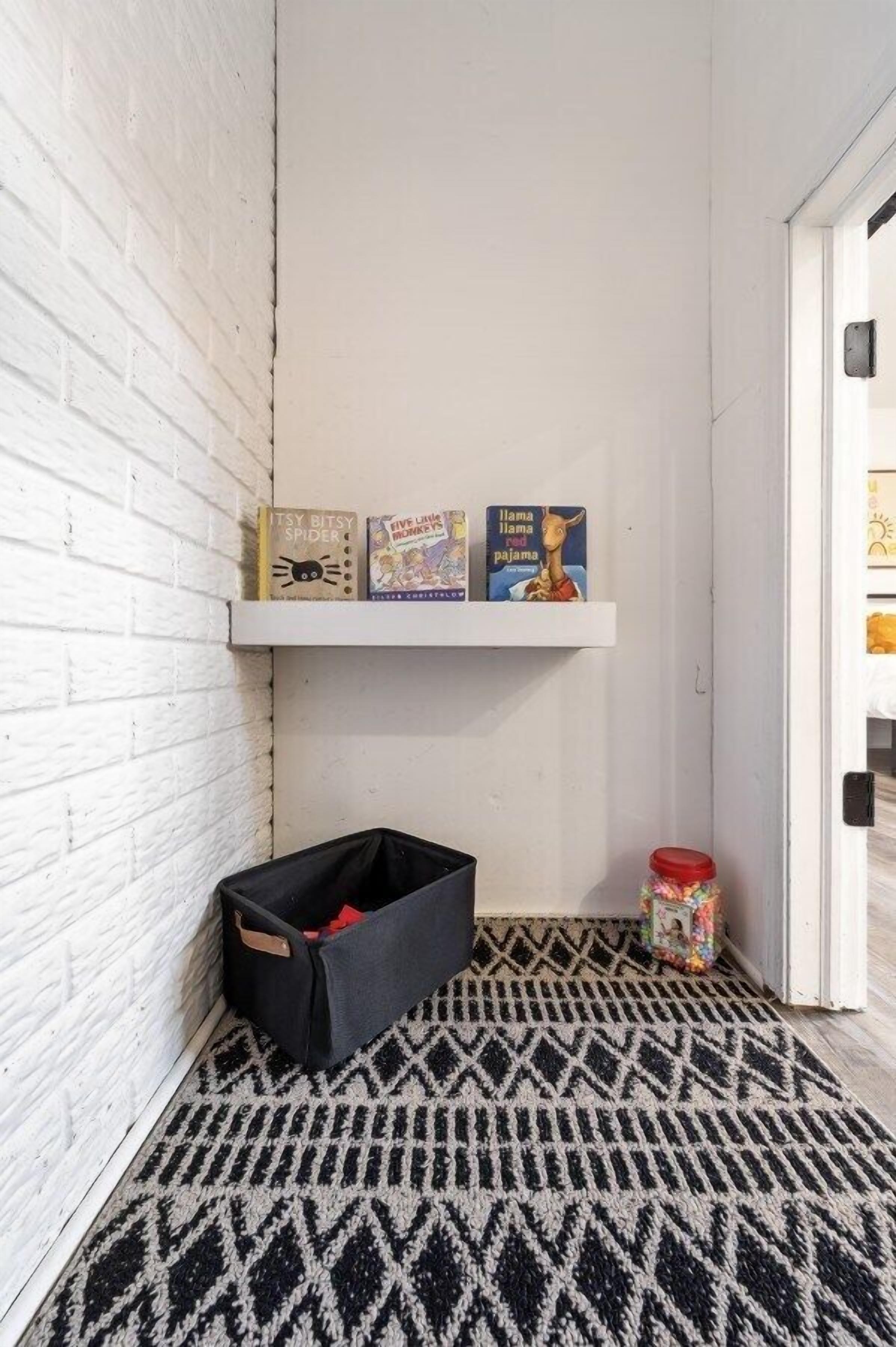 Una esquina con un muro de ladrillo blanco, un tapete de diseño blanco y negro, un cesto negro y una estantería con libros.