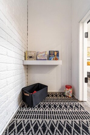 Een hoek met een witte bakstenen muur, een zwart-wit gestreept vloerkleed, een zwarte opbergbox en een plank met boeken.