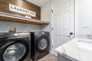 A laundry room with a Samsung washer and dryer, a wooden shelf with a sign, and a white door. - Huge Home, Gym, Game Room, Spa, BBQ, Near I-35 (Ankeny)