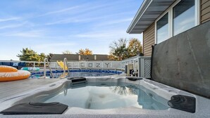 A hot tub with a wooden deck and a pool in the background.