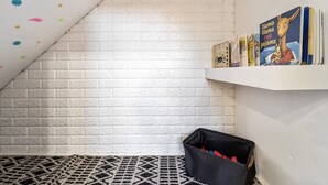 Une pièce avec un plafond en pente, un mur en briques blanches, un tapis à motifs noir et blanc, un bac de rangement noir et une étagère remplie de livres.