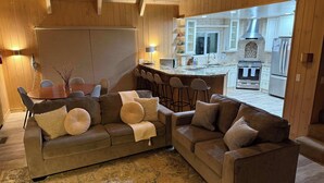 Un salon chaleureux avec un canapé en cuir marron, une table à manger et un coin cuisine visible à travers une porte ouverte.