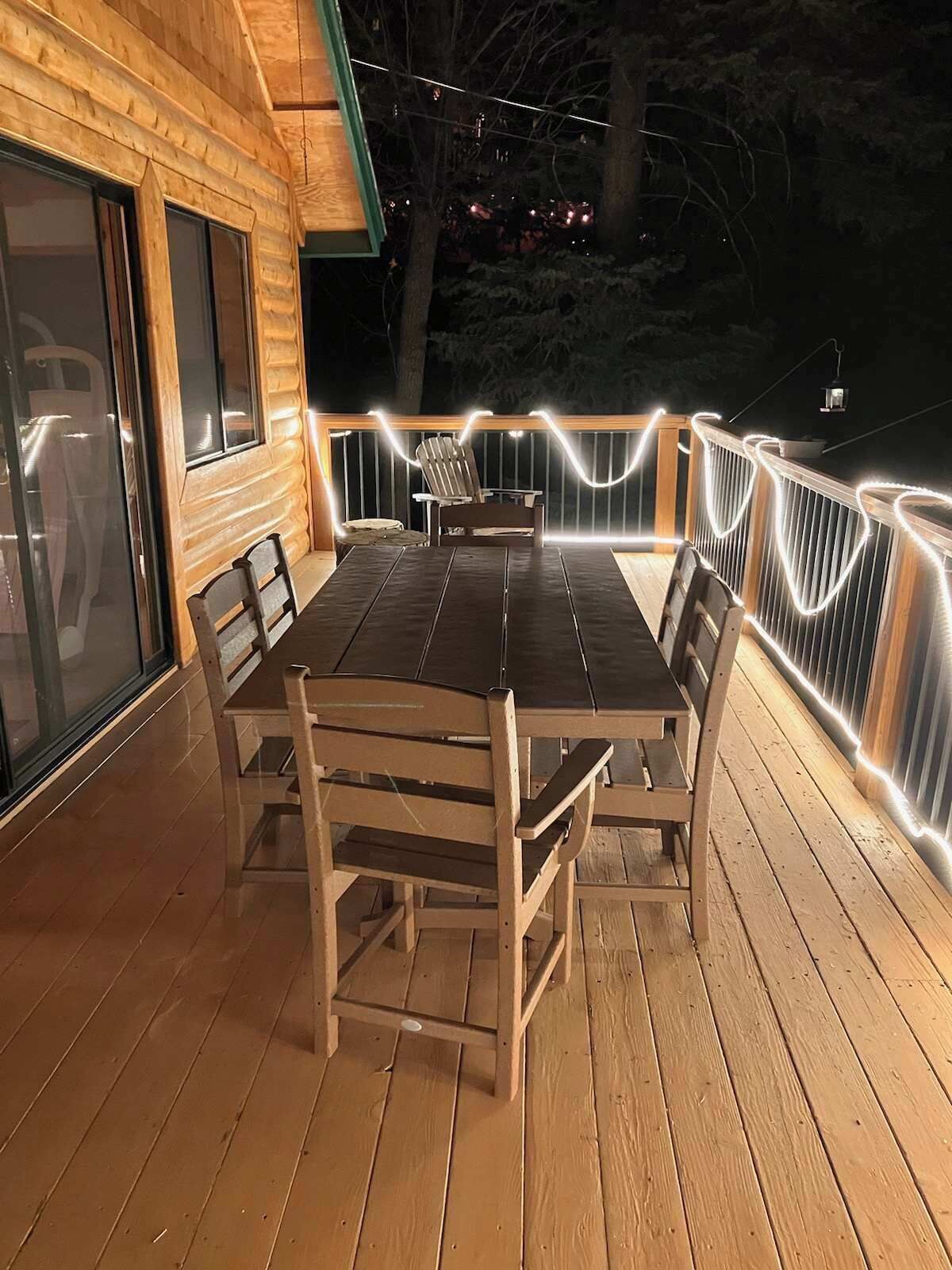 A wooden deck with a table and chairs, surrounded by a railing with string lights.