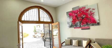 A reception area with a wooden archway leading to the outside, a grey sofa with cushions, and a large painting of a tree on the wall.