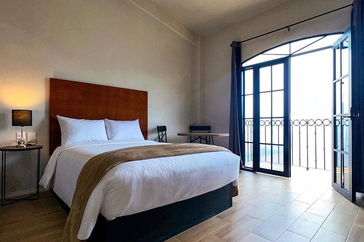A hotel room with a large bed, a nightstand with a lamp, a small table, and a balcony with a metal railing.