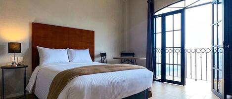 Una habitación de hotel con una cama grande, una mesita de noche con lámpara, una pequeña mesa y un balcón con barandilla de metal.