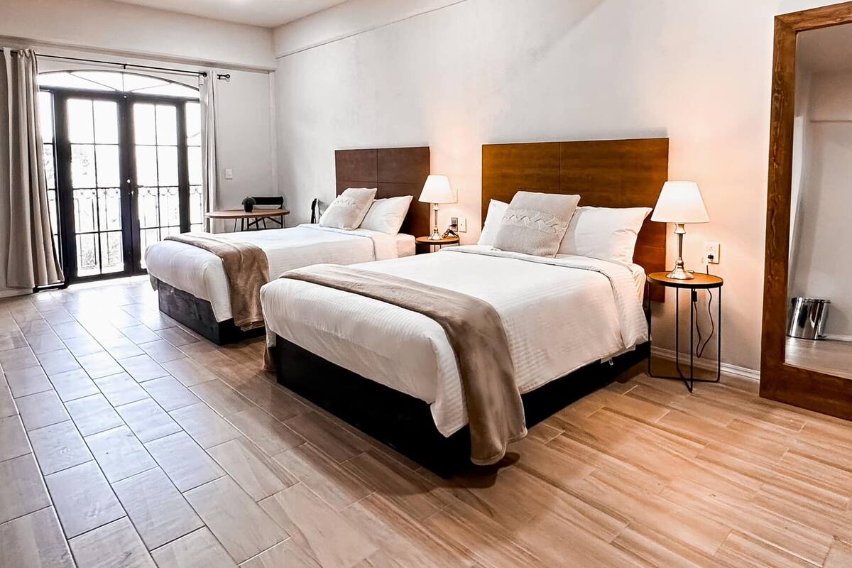 A hotel room with two beds, a wooden floor, and a large window with curtains.